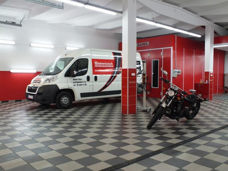 Warsztat tapicerski z białą furgonetką z logo firmy Szewczyk i motocyklem na szachownicowej podłodze, na tle czerwonej kabiny lakierniczej.
