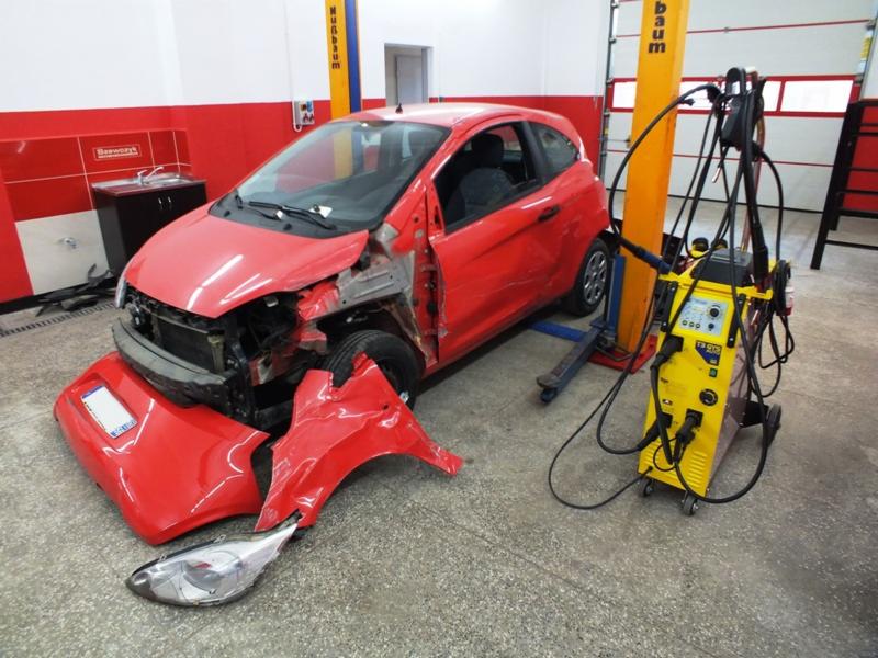 Czerwony samochód po wypadku w warsztacie, z uszkodzonym przodem, zderzak, błotnik i reflektor leżą na podłodze obok auta, obok stoi żółty sprzęt spawalniczy.