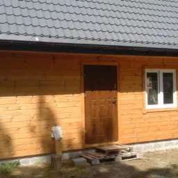 Niewielki, drewniany budynek z szarą blachodachówką, widoczne detale wykończenia, takie jak rynny i okno PCV, oraz prowizoryczne schody z palet.