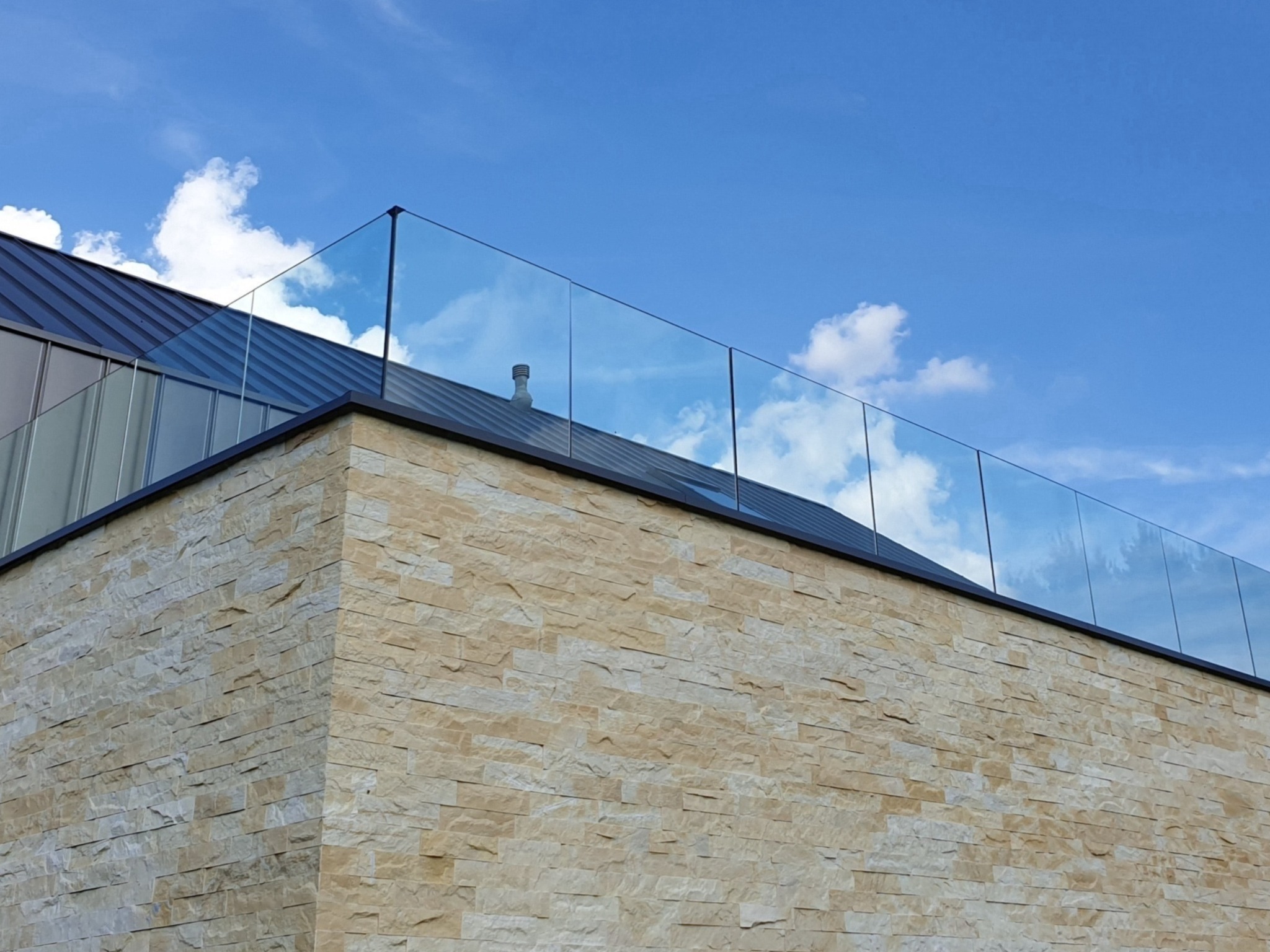 Nowoczesna elewacja budynku z kamienia z przezroczystą balustradą na dachu, błękitne niebo w tle.