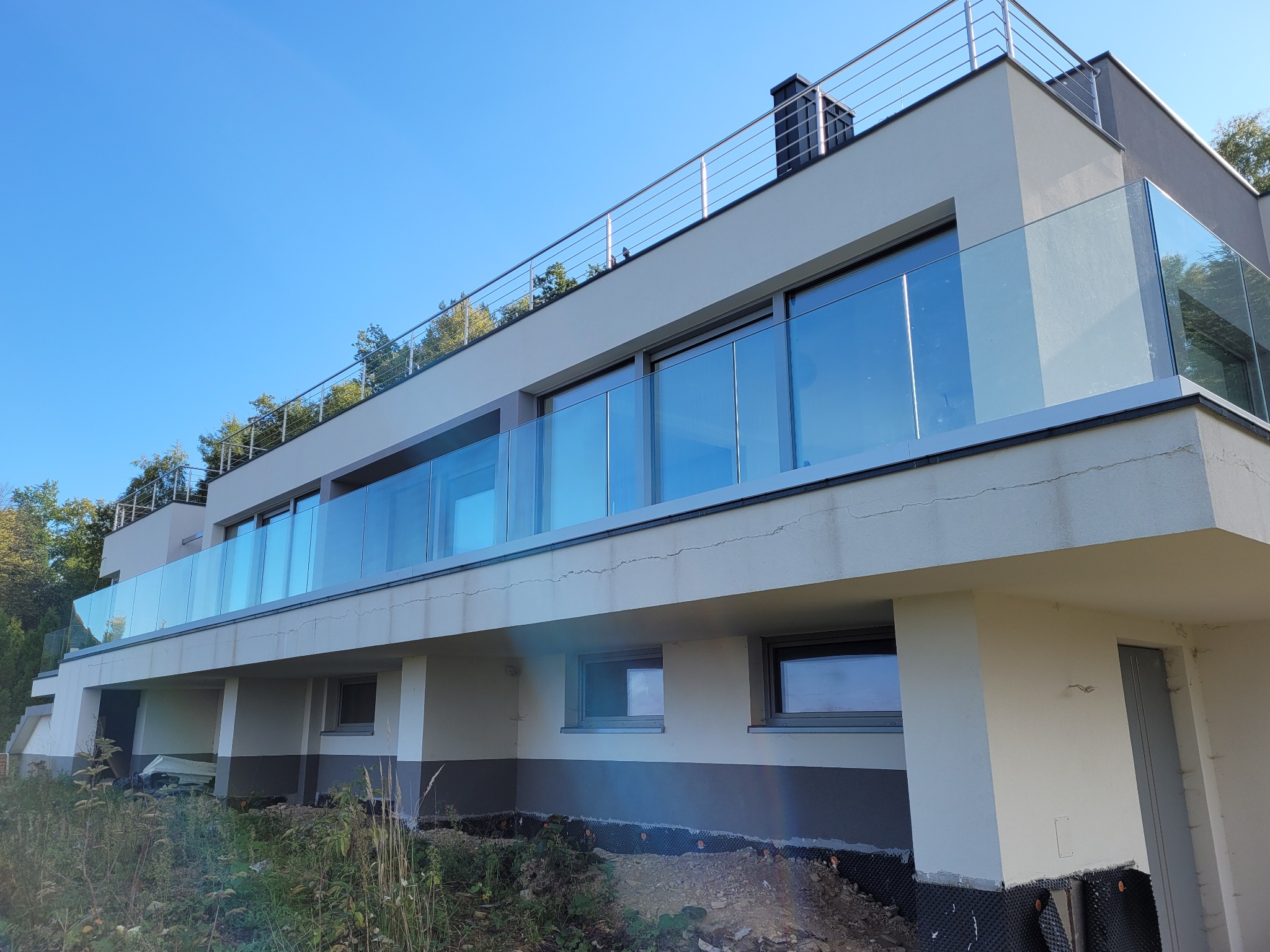 Nowoczesny budynek z balkonem zabezpieczonym szklaną taflą i metalową poręczą, widok z dołu na tle błękitnego nieba.