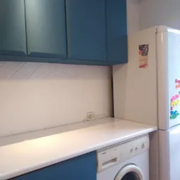 Niebieskie szafki kuchenne z białą płytką ułożoną w romby, biały blat, pralka i lodówka w tle.