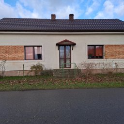 BEST-TRANS - Jednopiętrowy dom z kremową elewacją, brązowym dachem z blachodachówki i ceglanymi akcentami wokół okien, widziany z poziomu ulicy w pochmurny dzień.