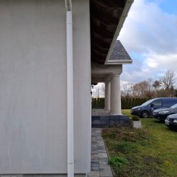 BEST-TRANS - Fragment elewacji domu z widoczną rynną spustową, kostką brukową na chodniku i kolumnami podtrzymującymi zadaszenie tarasu, w tle zaparkowane samochody.