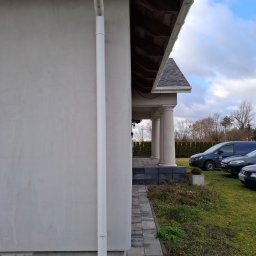 BEST-TRANS - Fragment elewacji domu z widoczną rynną spustową, obok ścieżka z kostki brukowej i podjazd z samochodami w tle.