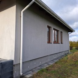 BEST-TRANS - Szara elewacja domu jednorodzinnego z brązowymi oknami i białym systemem rynnowym, widok z boku na fragment ściany z widoczną podmurówką i fragmentem chodnika z kostki.