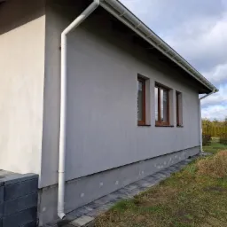 Szara elewacja domu jednorodzinnego z brązowymi oknami i białym systemem rynnowym, widok z boku na fragment ściany z widoczną podmurówką i fragmentem chodnika z kostki.