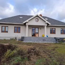 Nowo wybudowany dom z szarą elewacją, ciemną dachówką i schodami z kostki brukowej prowadzącymi do wejścia, otoczony niezagospodarowanym terenem.