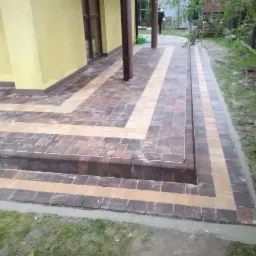 Ułożona kostka brukowa w geometryczny wzór przed domem, z widocznymi schodami i obrzeżami, w tle żółta elewacja i osoba z konewką.