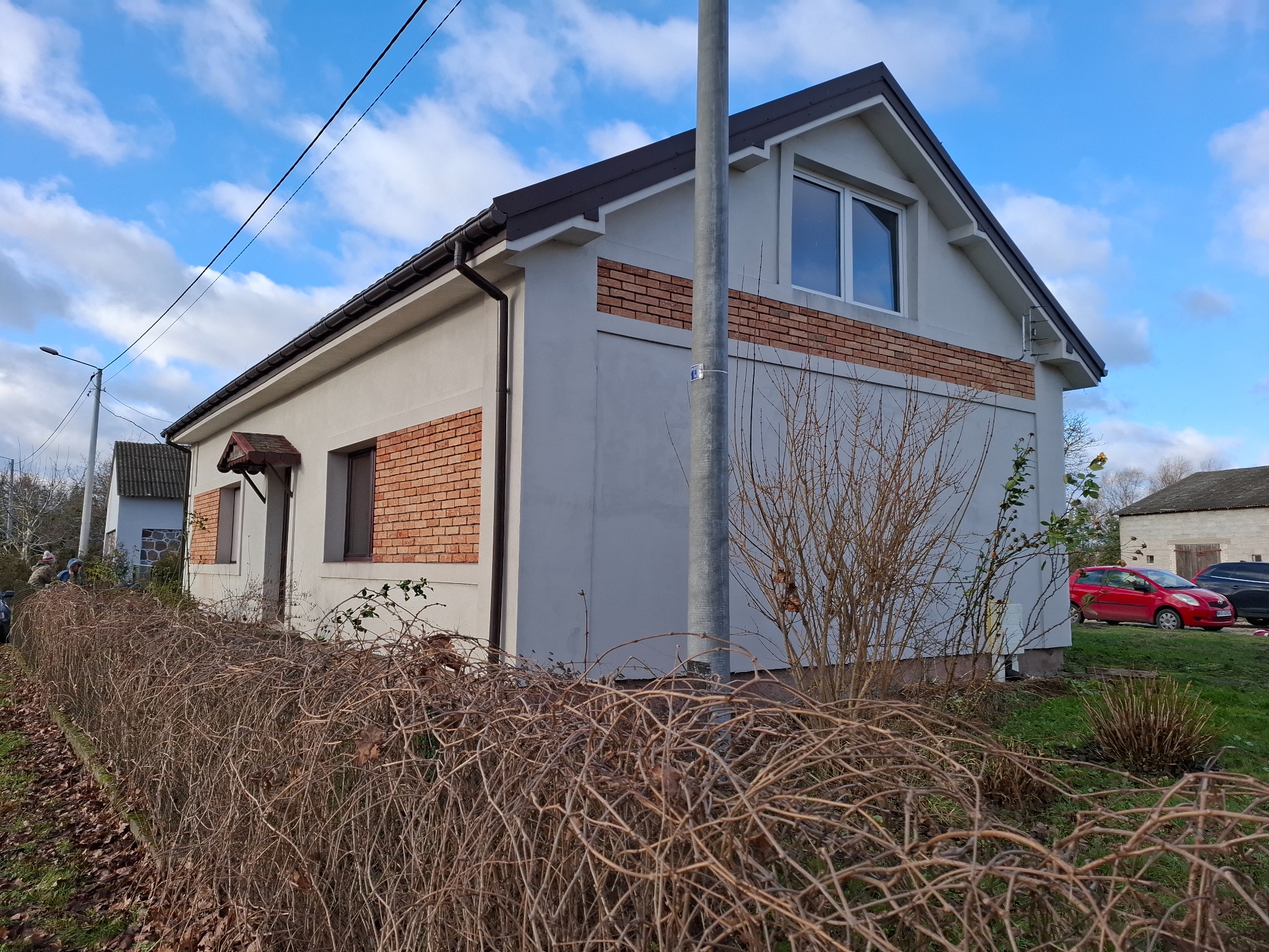 Dwukondygnacyjny dom z szarą elewacją i paskami z czerwonej cegły, brązowy dach, widok zza niskiego żywopłotu, błękitne niebo w tle.