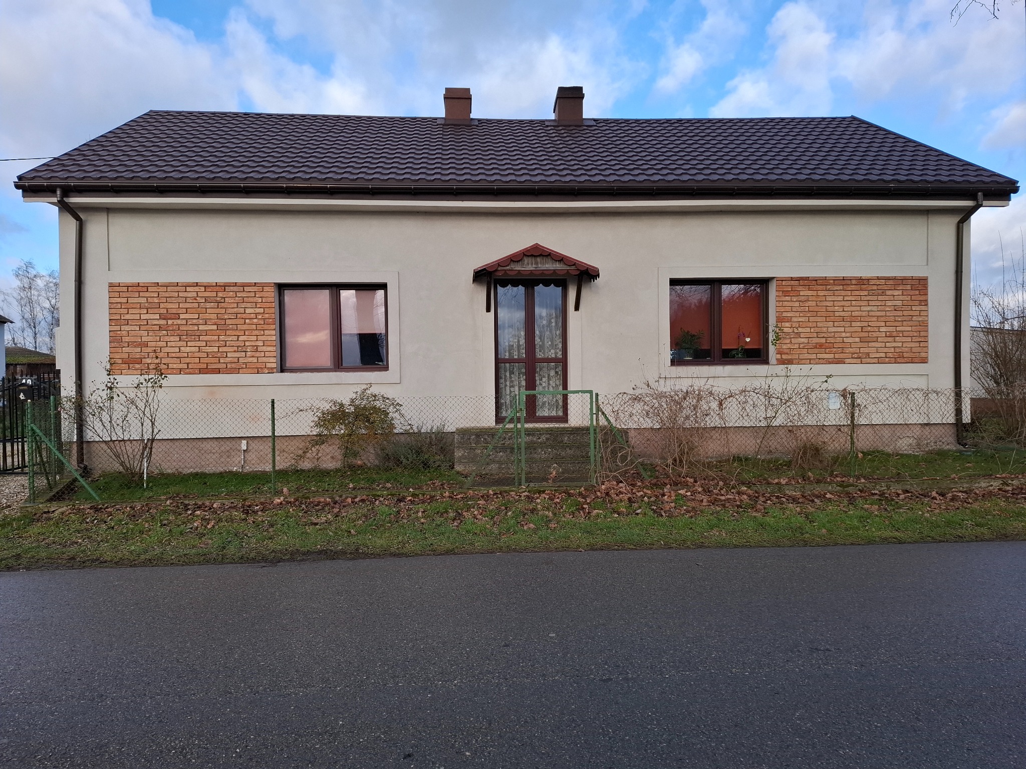 Jednopiętrowy dom z kremową elewacją, brązowym dachem z blachodachówki i ceglanymi akcentami wokół okien, widziany z poziomu ulicy w pochmurny dzień.