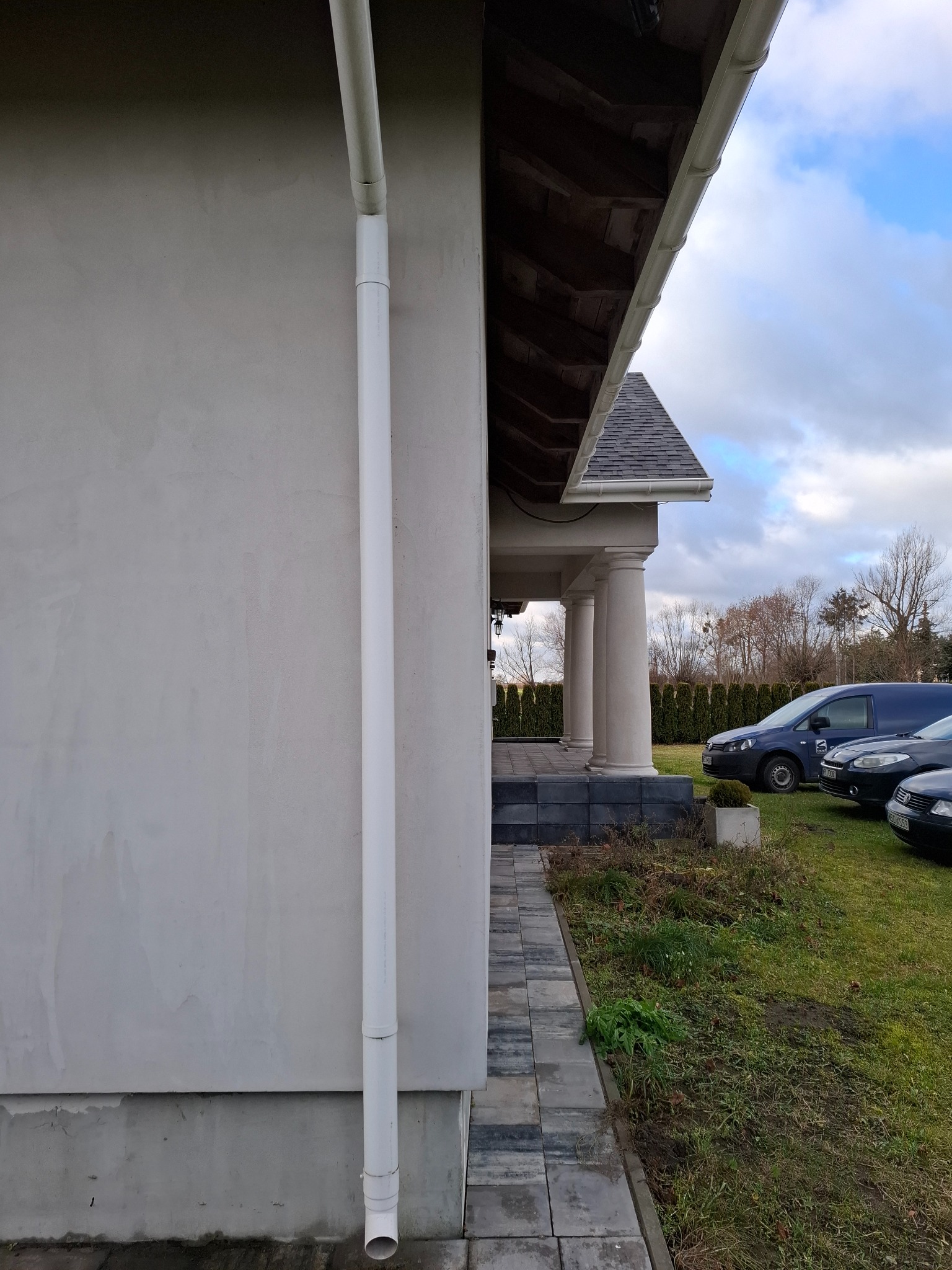 Fragment elewacji domu z widoczną rynną spustową, kostką brukową na chodniku i kolumnami podtrzymującymi zadaszenie tarasu, w tle zaparkowane samochody.