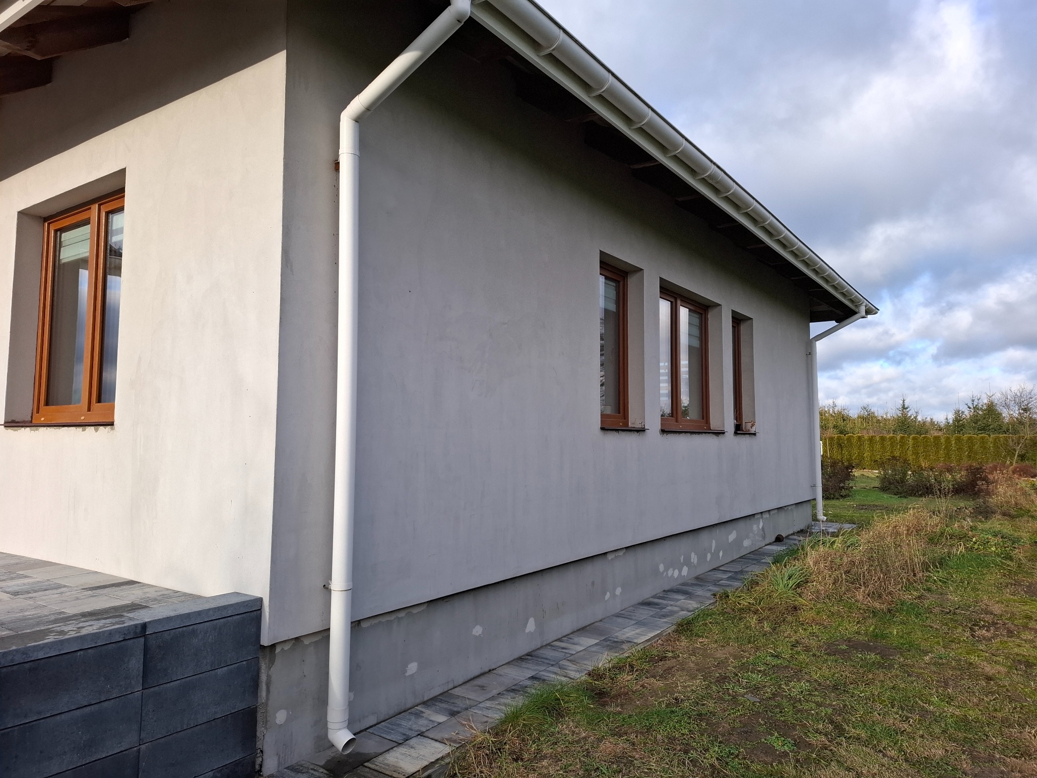 Szara elewacja domu jednorodzinnego z brązowymi oknami i białym systemem rynnowym, widok z boku na fragment ściany z widoczną podmurówką i fragmentem chodnika z kostki.