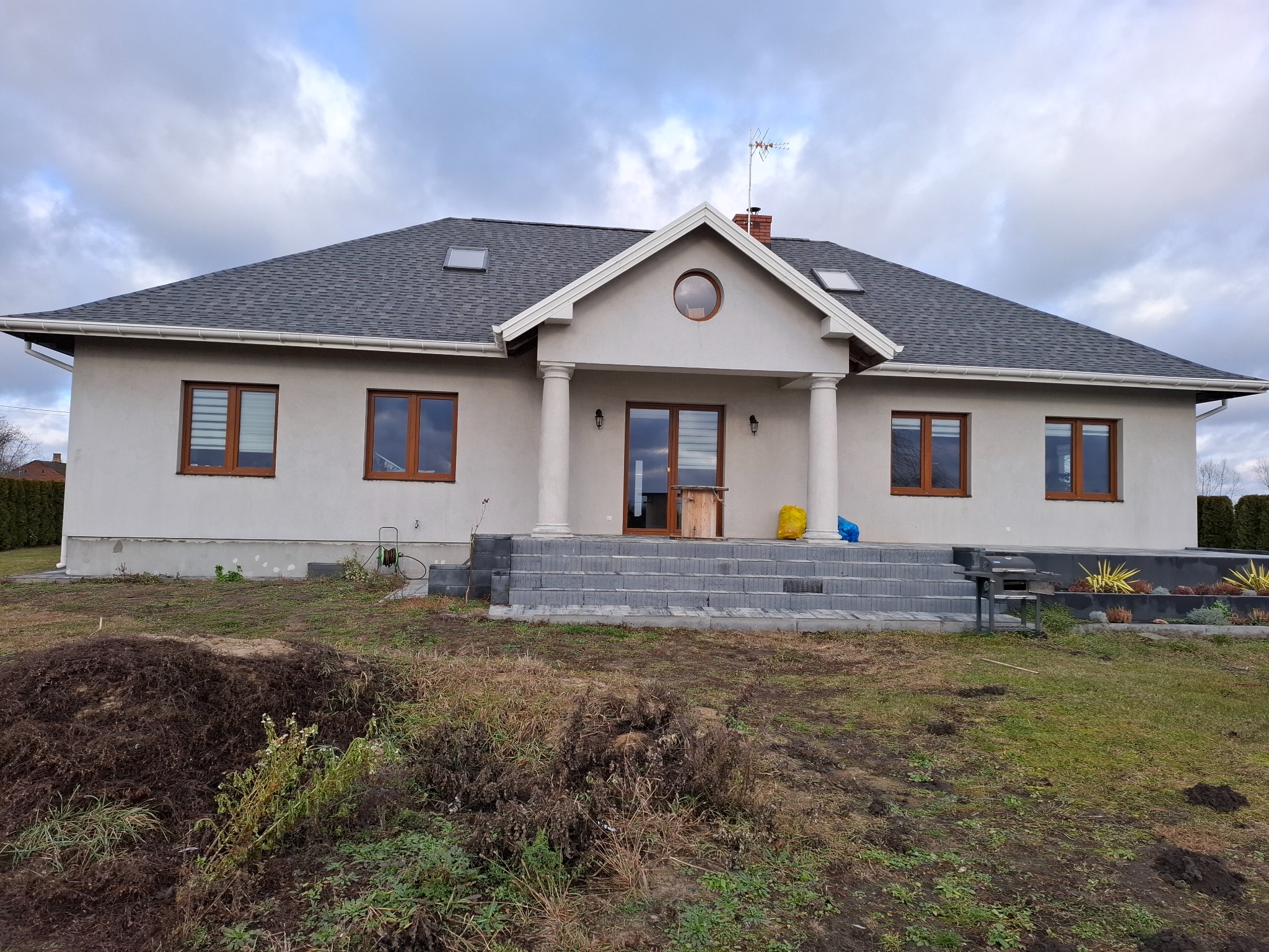 Nowo wybudowany dom z szarą elewacją, ciemną dachówką i schodami z kostki brukowej prowadzącymi do wejścia, otoczony niezagospodarowanym terenem.