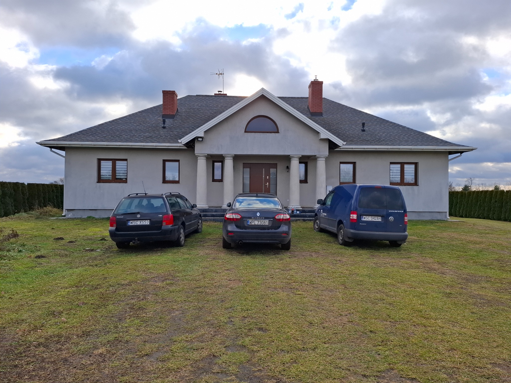 Parterowy dom z szarą elewacją, kolumnami przy wejściu i trzema samochodami zaparkowanymi na trawniku przed nim, pod pochmurnym niebem.