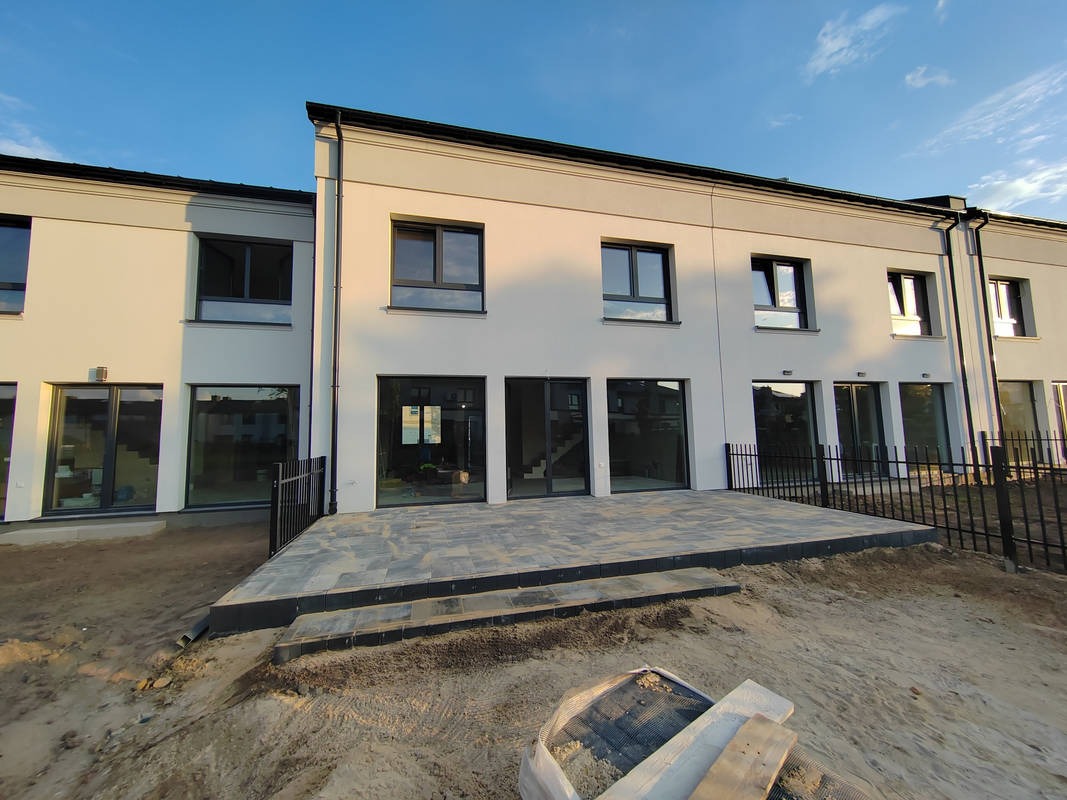 Nowoczesny szeregowiec z szarą elewacją, czarnymi oknami i tarasem wyłożonym szarą kostką brukową. Widoczne przygotowania do zagospodarowania terenu wokół.