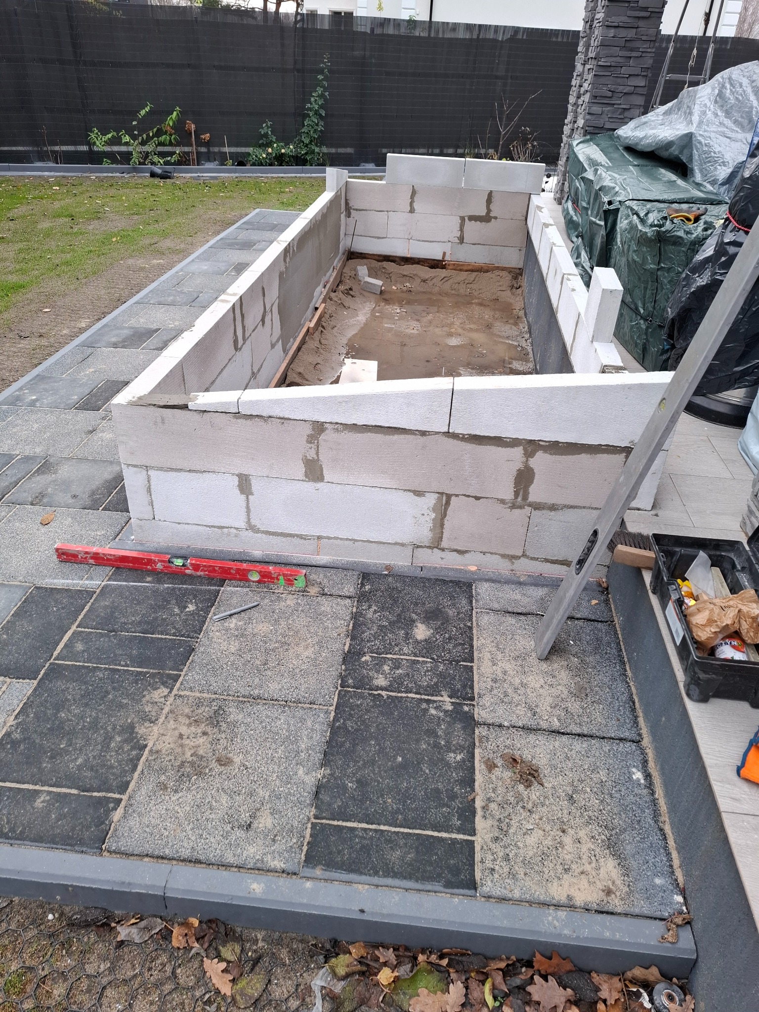 Wznoszenie murku z bloczków betonowych na posesji, widoczny poziomica, narzędzia w skrzynce i ułożona kostka brukowa dookoła