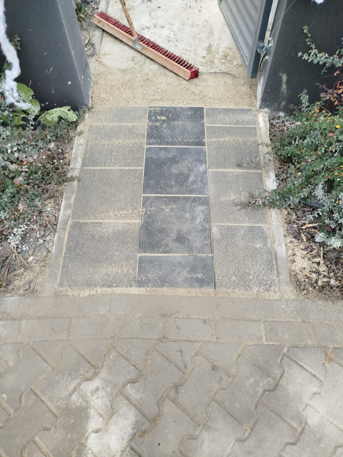 Ułożona kostka brukowa w odcieniach szarości, z ciemniejszym pasem centralnym, częściowo zasypana piaskiem, obok furtki i z widoczną szczotką do zamiatania.