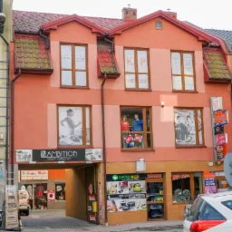 Fasada budynku z szyldami firm, w tym 'KP Biuro Rachunkowe' widoczne w oknach, oraz inne sklepy i usługi w parterze budynku.