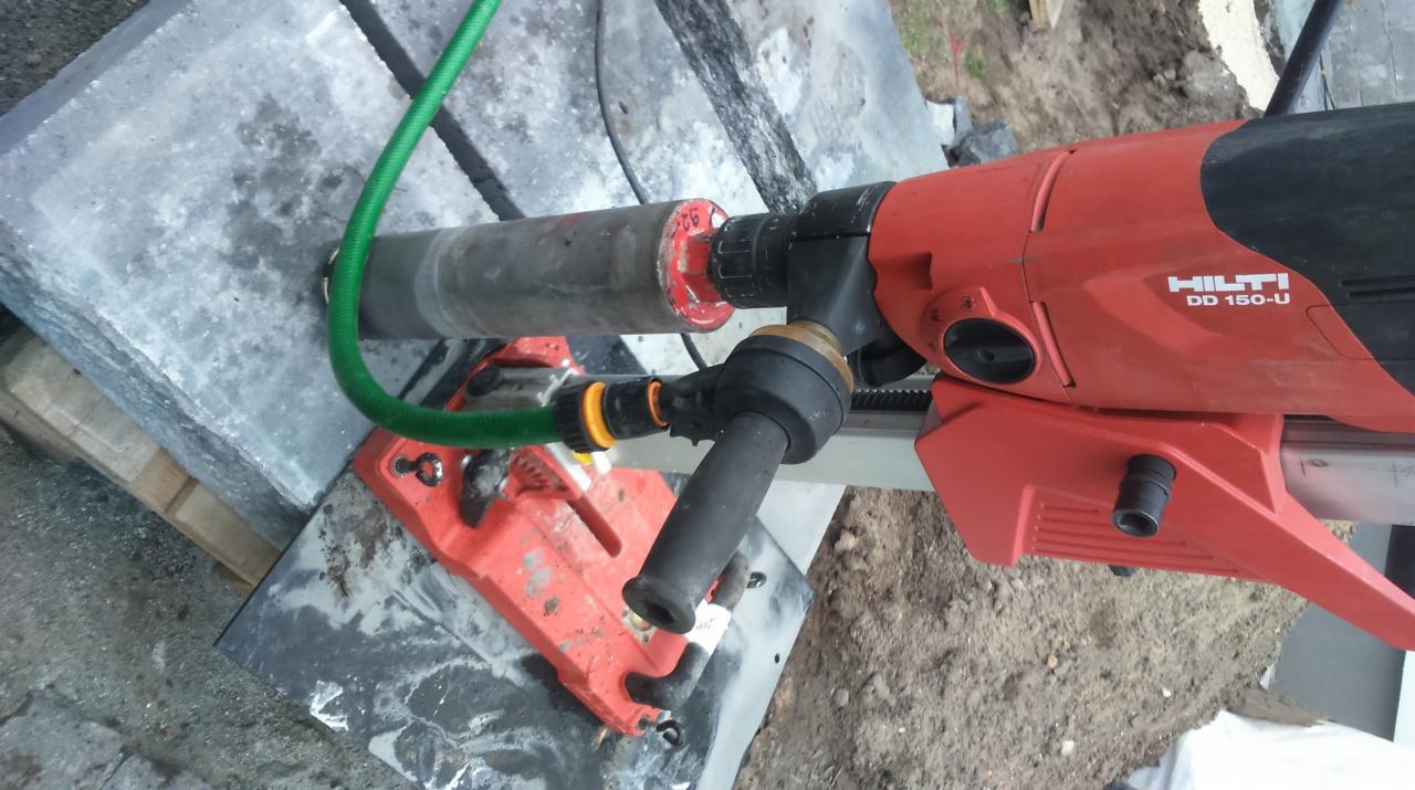 Wiertnica Hilti DD 150-U z wiertłem koronowym i podłączeniem do węża z wodą, wiercąca otwór w szarej płycie chodnikowej.