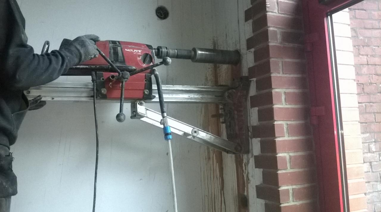 Czerwona wiertnica diamentowa Hilti na aluminiowej szynie, wiercąca otwór w murze z czerwonej cegły, widoczna ściana z białego betonu oraz częściowo osoba w roboczym ubraniu.