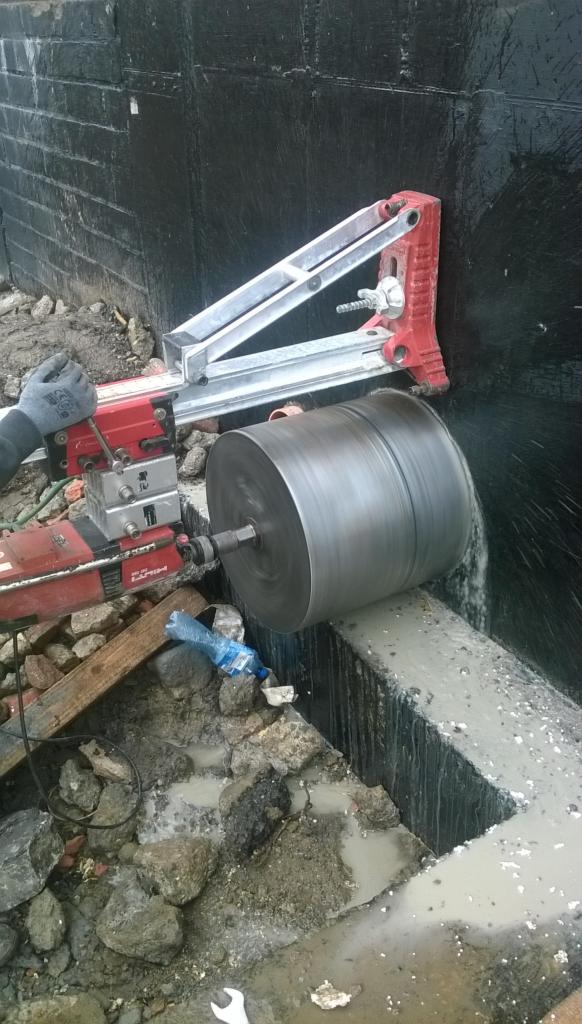 Wiercenie rdzeniowe w żelbetowej ścianie fundamentowej z użyciem wiertnicy Hilti, woda chłodząca spływa po ścianie, widoczne gruz i narzędzia.