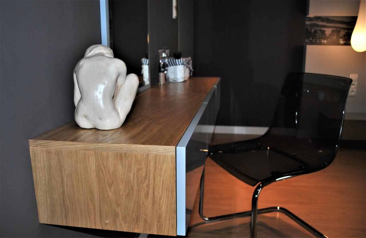 Nowoczesna toaletka z blatem z jasnego drewna, ozdobiona białą rzeźbą siedzącej postaci, obok krzesło z przezroczystego tworzywa na chromowanym stelażu, na tle ciemnoszarej ściany i drewnianej...