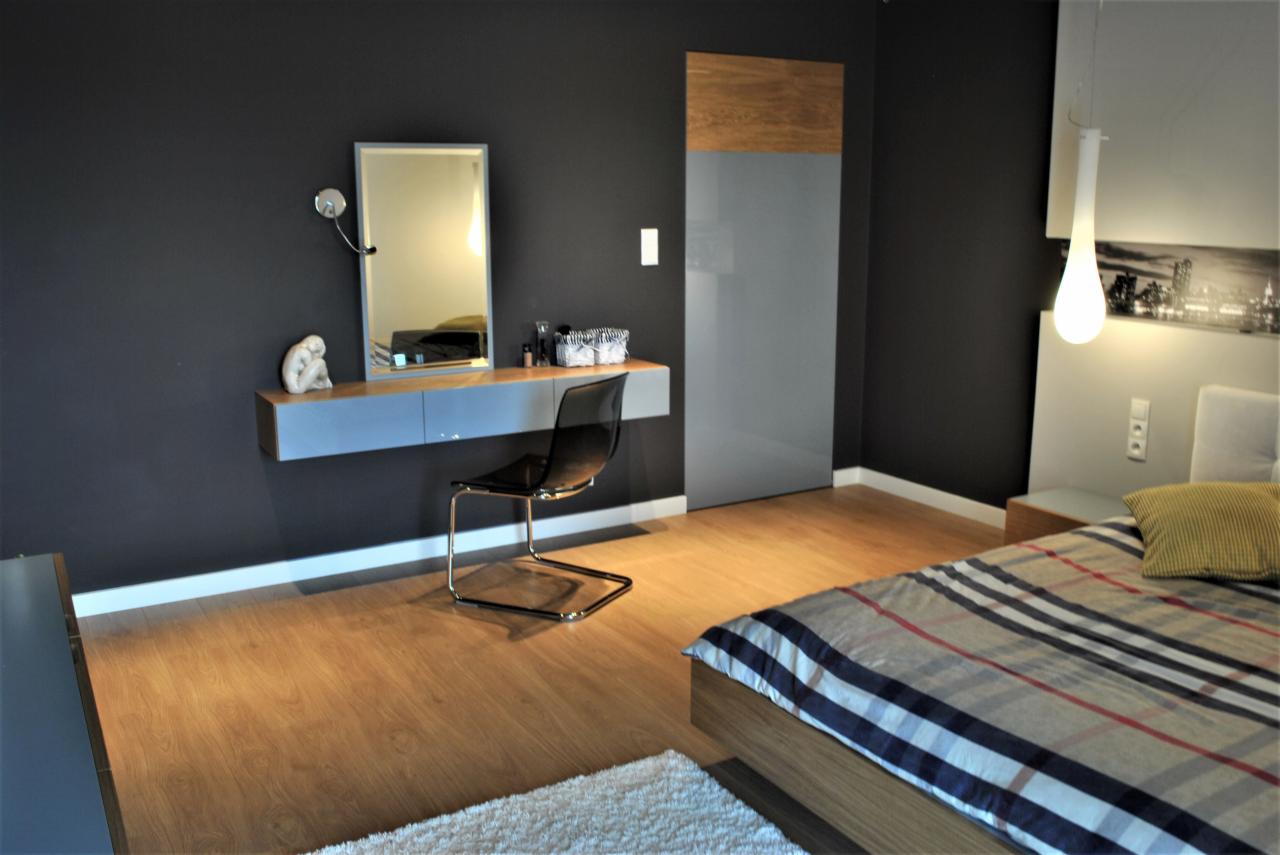 Nowoczesna sypialnia z toaletką na ścianie w kolorze grafitowym, przezroczyste krzesło, drewniana podłoga i dywanik z długim włosem.
