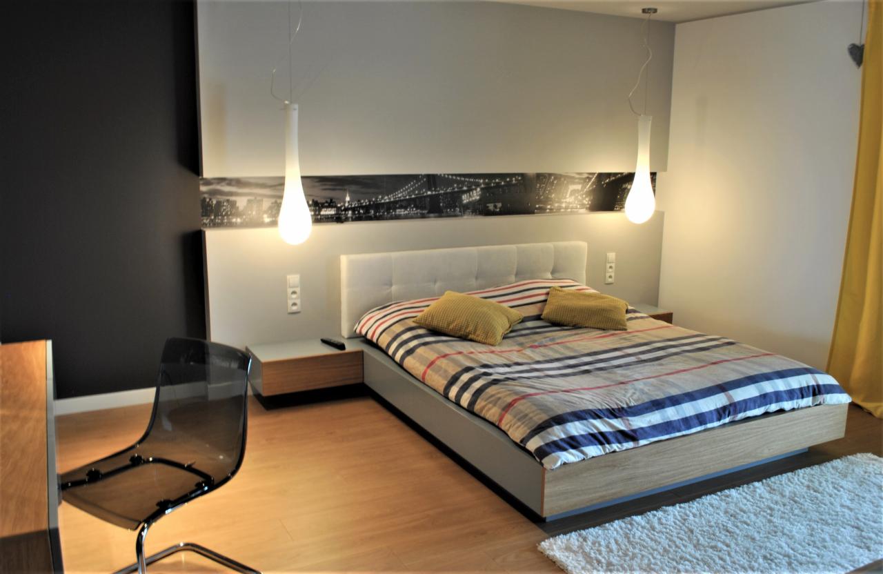 Nowoczesna sypialnia z łóżkiem w paski, podwieszanymi lampami w kształcie kropli i czarnobiałym pasem panoramy miasta nad wezgłowiem, uzupełniona przezroczystym krzesłem i puszystym dywanikiem.