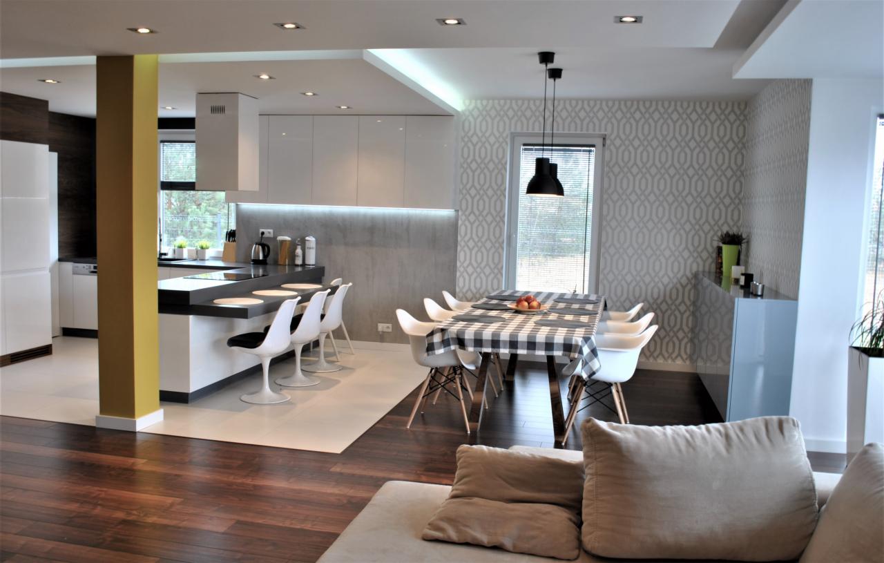 Nowoczesna kuchnia z wyspą i jadalnią, białe szafki, czarny blat, żółta kolumna, stół nakryty w czarno-białą kratę, białe krzesła, wzorzysta tapeta w odcieniach szarości, drewniana podłoga.