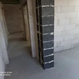 Wnętrze w trakcie budowy domu murowanego, widoczny fragment ściany z białych bloczków i czarny filar z betonu komórkowego na pierwszym planie.