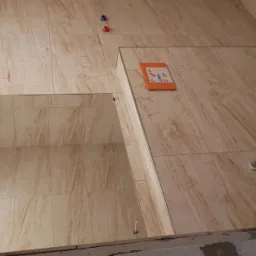 Łazienka w trakcie wykończenia: beżowe płytki imitujące marmur na ścianach, przygotowane otwory na armaturę, widoczne kable elektryczne, zabudowa wnęki z lustrem.