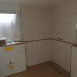 Wnętrze łazienki w trakcie prac wykończeniowych: białe płytki ścienne z ciemnoszarą fugą, widoczne przyłącza wodne, odpływ liniowy w podłodze, okno z pudełkiem i folią ochronną na parapecie...