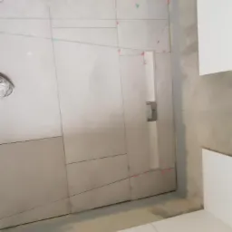 Ściana z szarych płytek, w trakcie układania, z widocznymi liniami pomocniczymi, krzyżykami dystansowymi i fragmentem poziomicy przy suficie, w tle białe płytki podłogowe i fragment białej szafki.