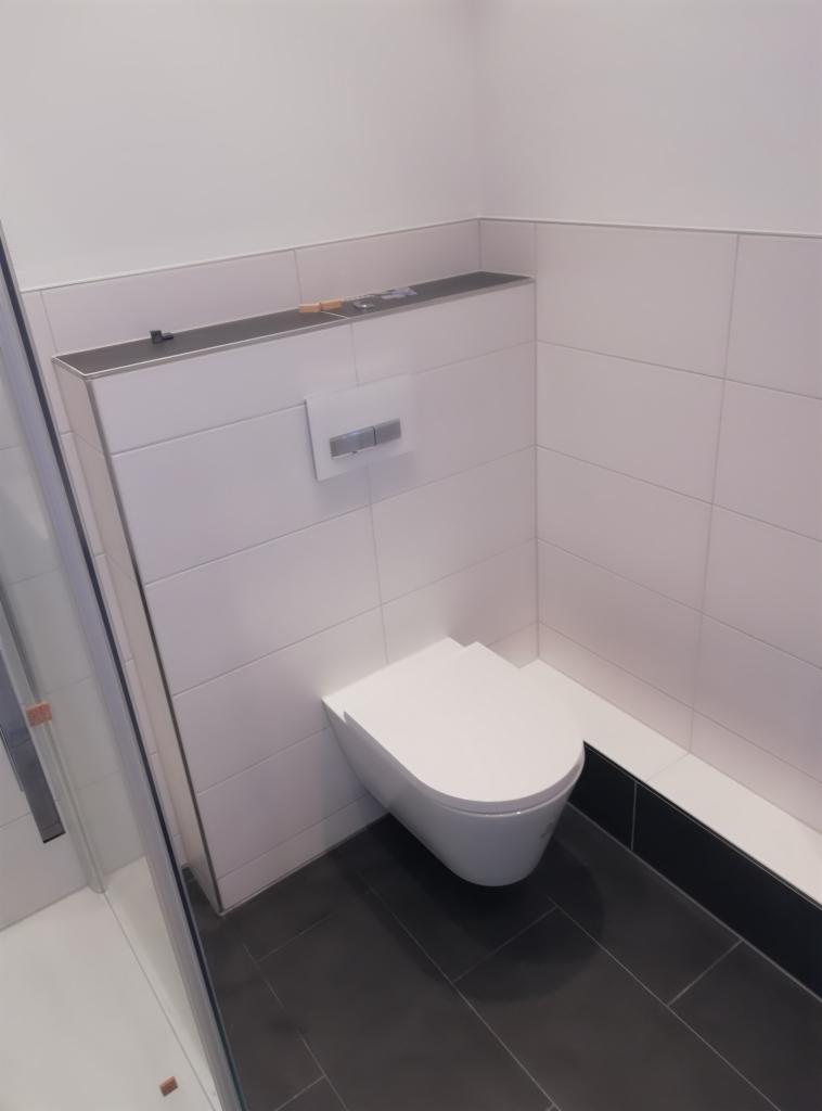 Nowoczesna toaleta podwieszana z białymi płytkami ściennymi i ciemną podłogą, widoczny fragment kabiny prysznicowej z boku.