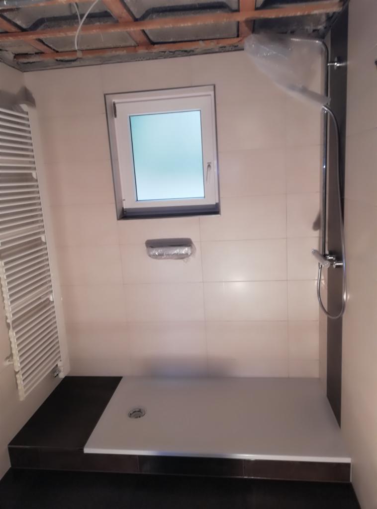 Niedokończona łazienka z jasnymi płytkami, brodzikiem z odpływem, grzejnikiem drabinkowym i panelem prysznicowym; surowy sufit z widocznymi belkami.