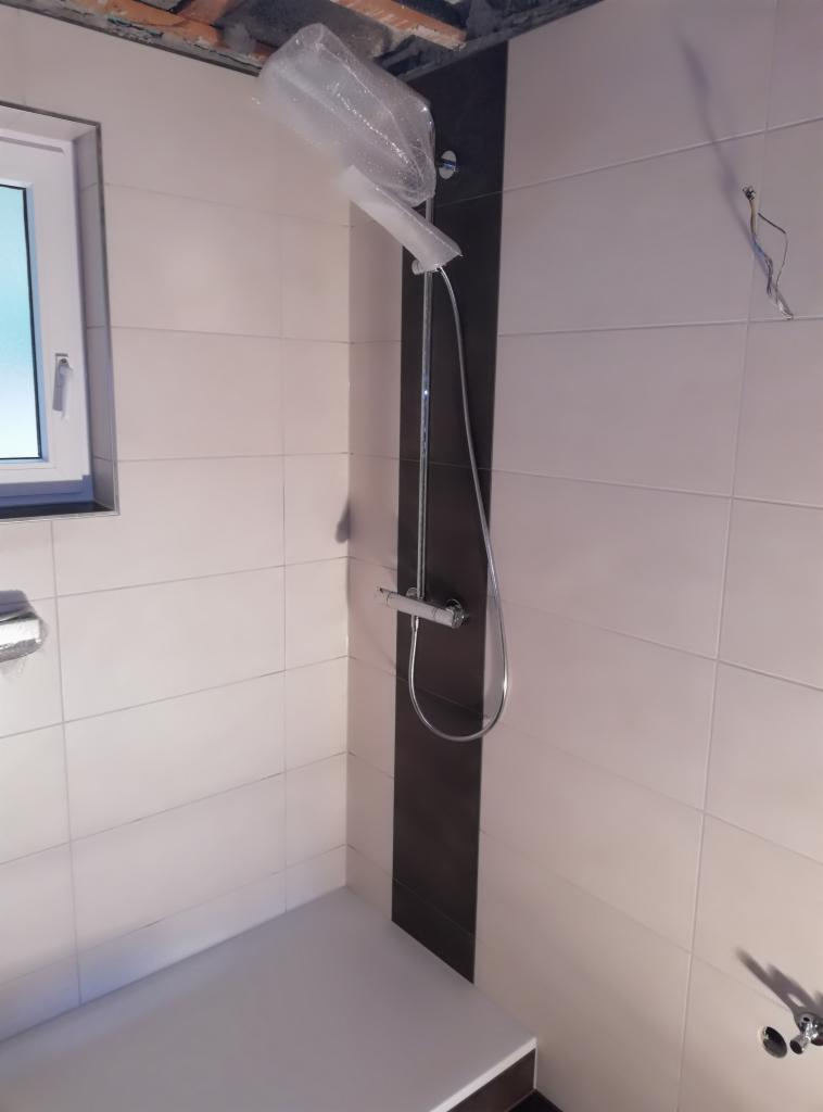 Niedokończona łazienka z białymi i brązowymi płytkami, zamontowanym prysznicem z folią ochronną na deszczownicy i widoczną surową konstrukcją sufitu.