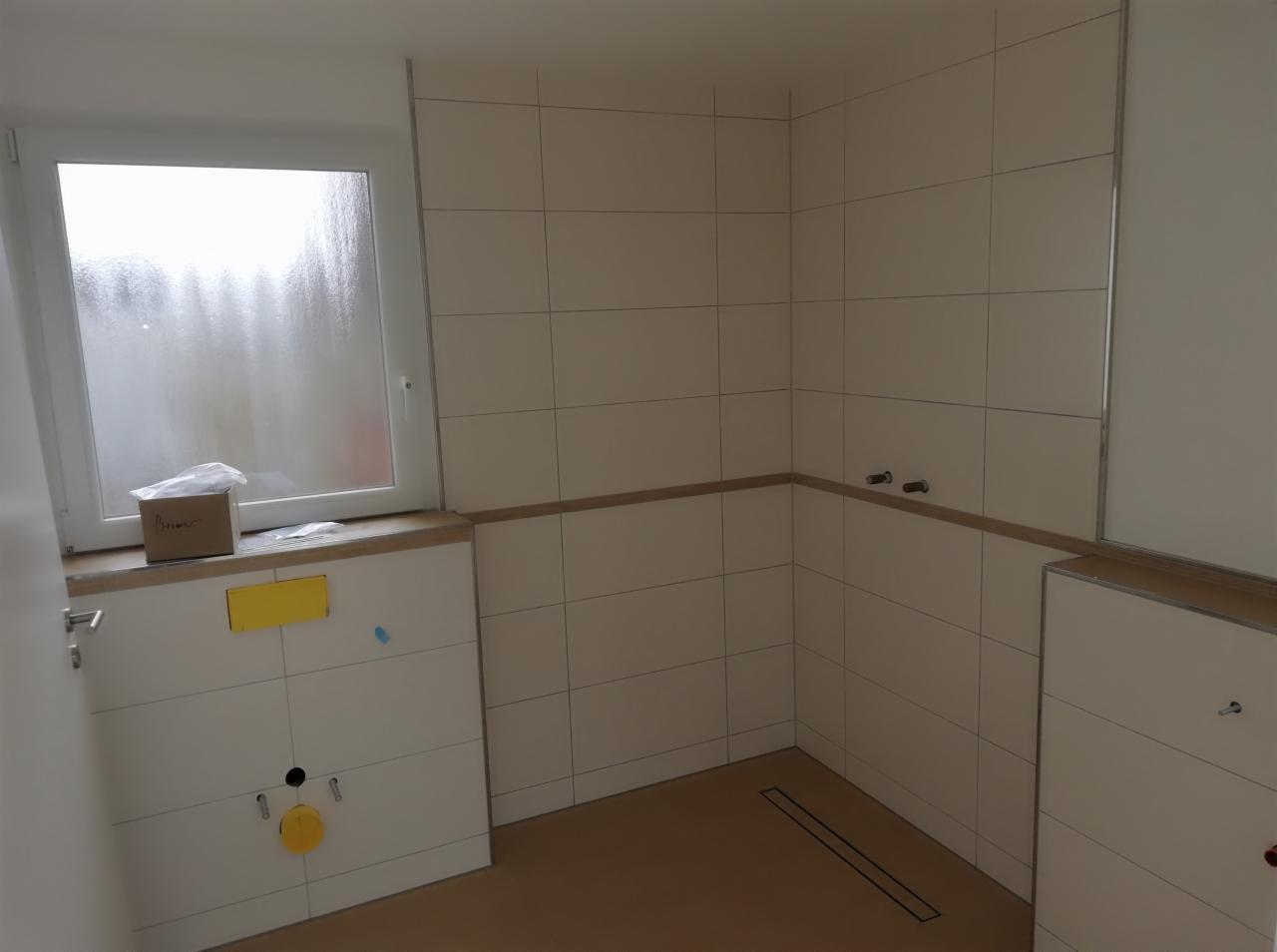 Wnętrze łazienki w trakcie prac wykończeniowych: białe płytki ścienne z ciemnoszarą fugą, widoczne przyłącza wodne, odpływ liniowy w podłodze, okno z pudełkiem i folią ochronną na parapecie...