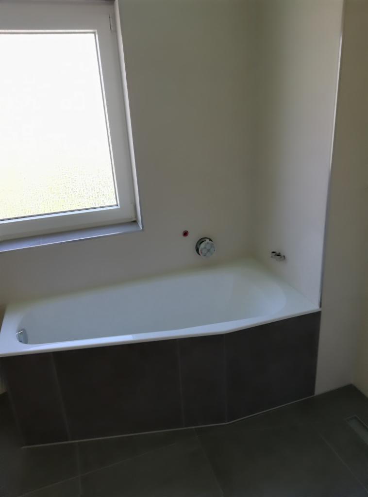 Biała wanna obudowana ciemnoszarymi płytkami w łazience z oknem z matową szybą. Widoczny fragment podłogi z szarych płytek i odpływ liniowy.