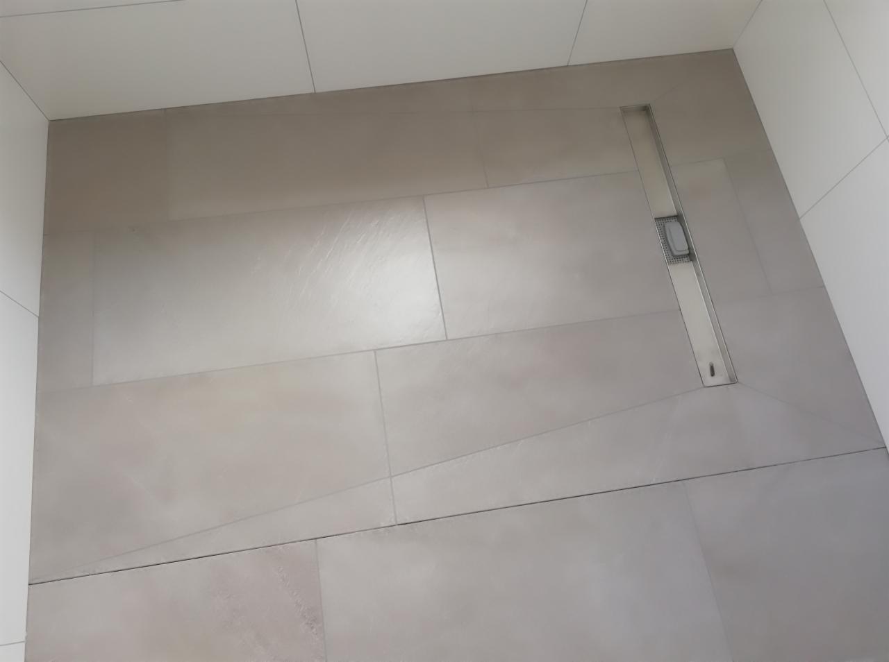 Beżowe, prostokątne płytki podłogowe w kabinie prysznicowej z odpływem liniowym ze stali nierdzewnej, widoczne fugi i białe płytki na ścianach.