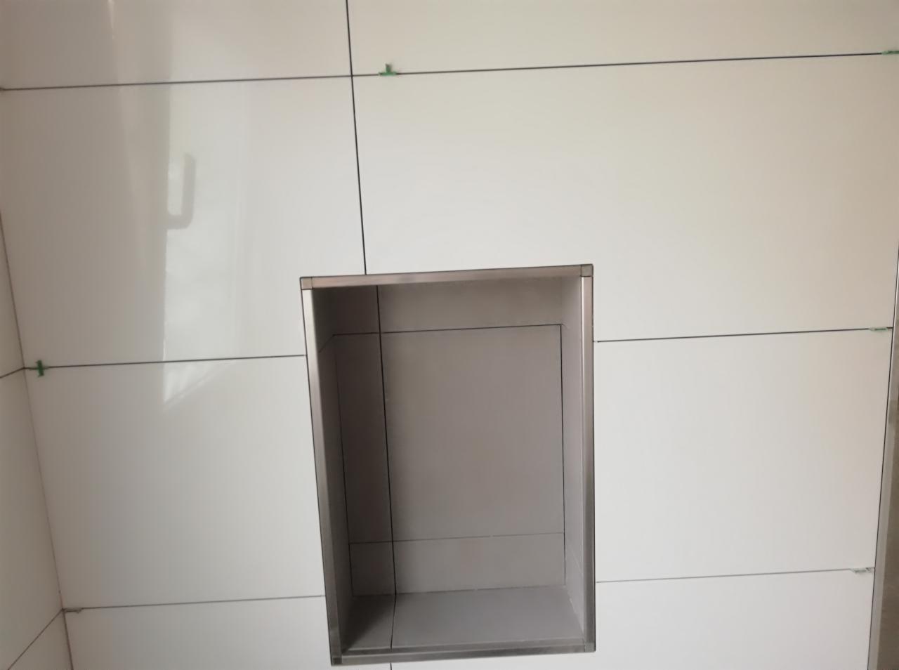 Białe, błyszczące płytki ścienne w łazience z wnęką wykończoną metalową ramką, widoczne małe, zielone krzyżyki dystansowe.