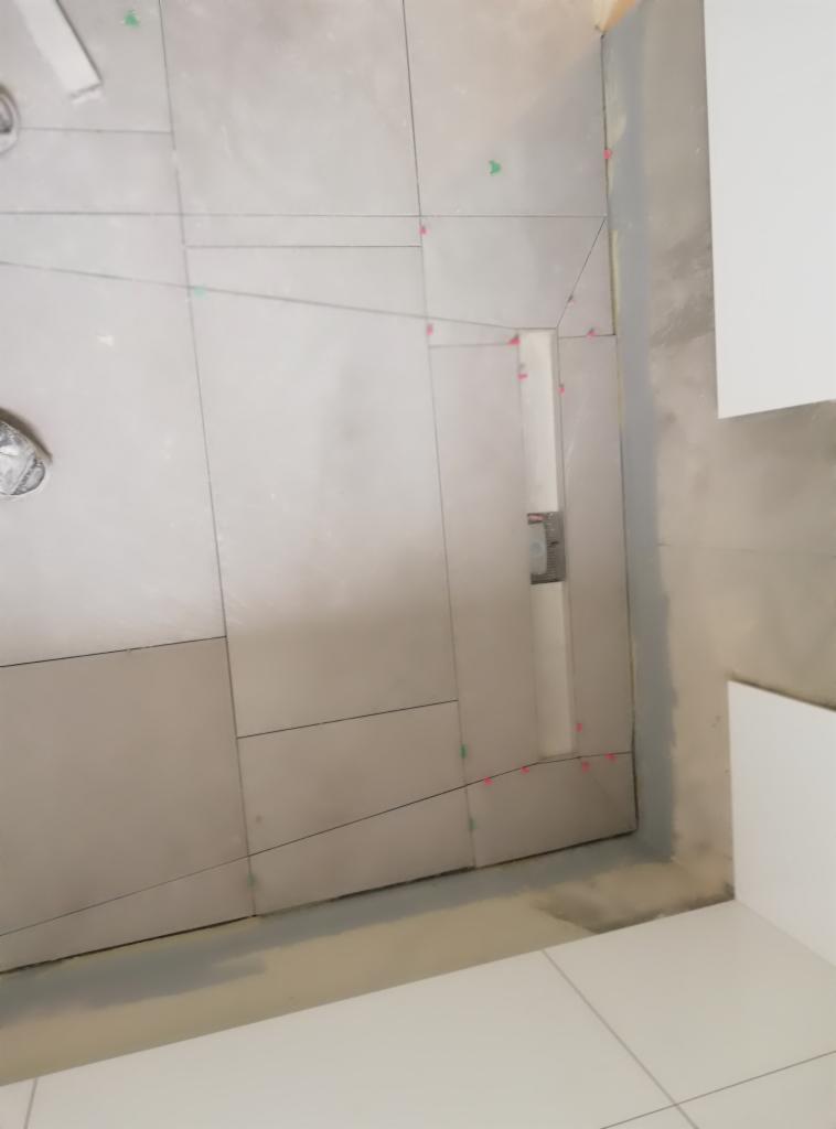 Ściana z szarych płytek, w trakcie układania, z widocznymi liniami pomocniczymi, krzyżykami dystansowymi i fragmentem poziomicy przy suficie, w tle białe płytki podłogowe i fragment białej szafki.
