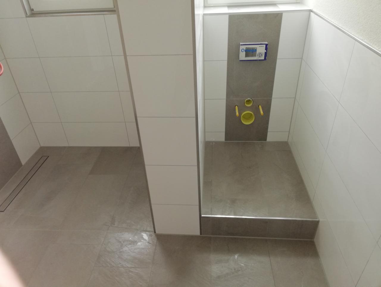 Wykończona strefa prysznicowa z odpływem liniowym i podtynkową instalacją WC na tle białych i szarych płytek ceramicznych.