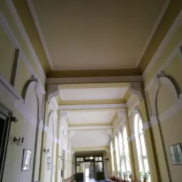 Długi korytarz w budynku z kremowymi ścianami, białymi detalami architektonicznymi i rzędami okien po prawej stronie, oświetlony naturalnym światłem.