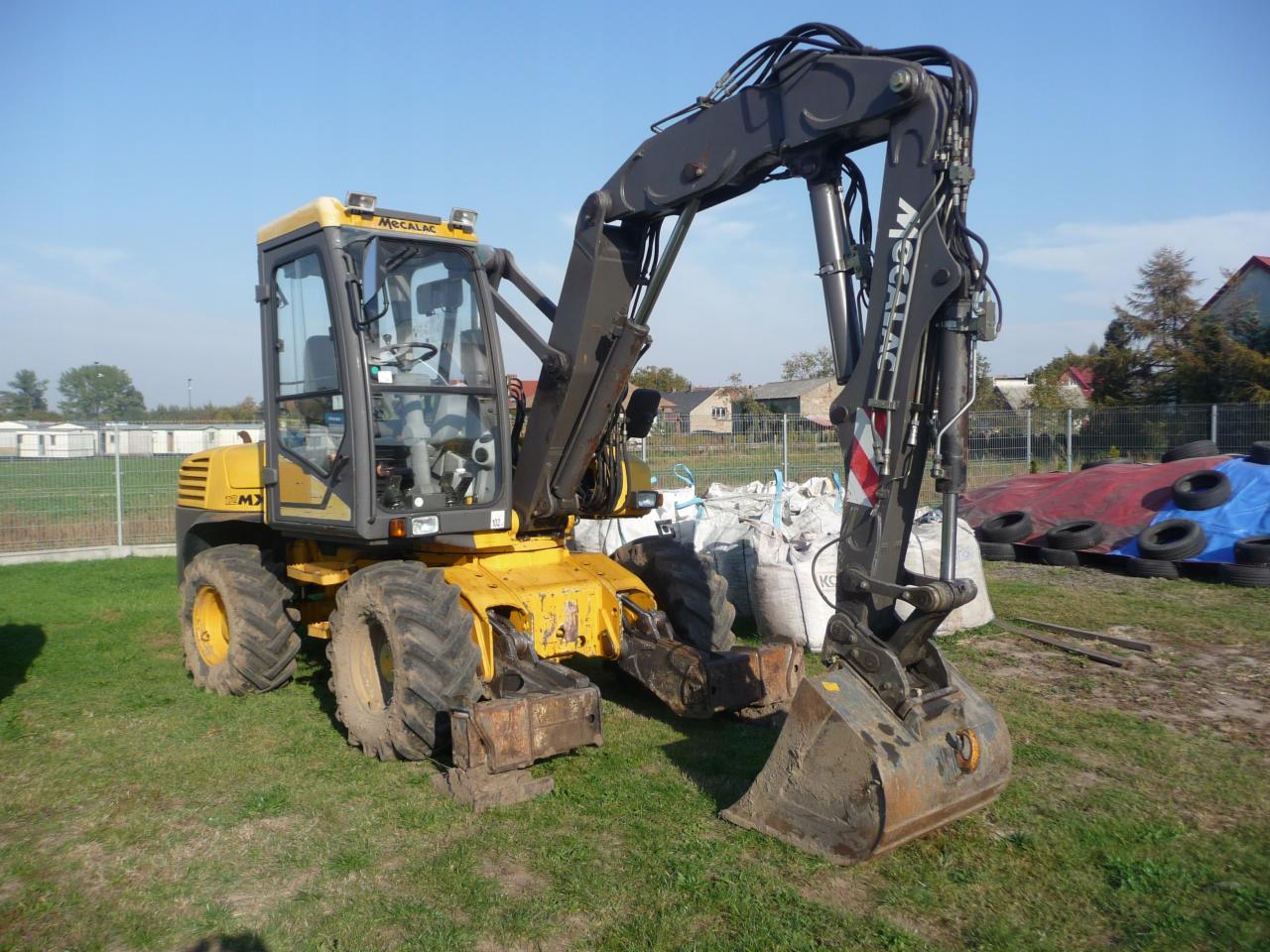Żółto-czarna koparka Mecalac na trawiastym terenie, z widocznymi workami z ziemią i oponami w tle, pod błękitnym niebem.