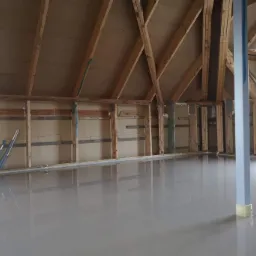 Świeżo wylana, gładka posadzka w surowym wnętrzu poddasza z widoczną konstrukcją drewnianą i stalową podporą.