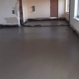 Świeżo wylana, gładka, szara posadzka w przestronnym, remontowanym wnętrzu. Folia ochronna przy ścianach, przygotowanie do dalszych prac wykończeniowych.