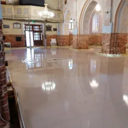 Wnętrze kościoła z nową, jasną wylewką na podłodze, odbijającą światło żyrandola i łuki architektoniczne, z widocznymi fragmentami ceglanych filarów i przygotowanymi obrzeżami.