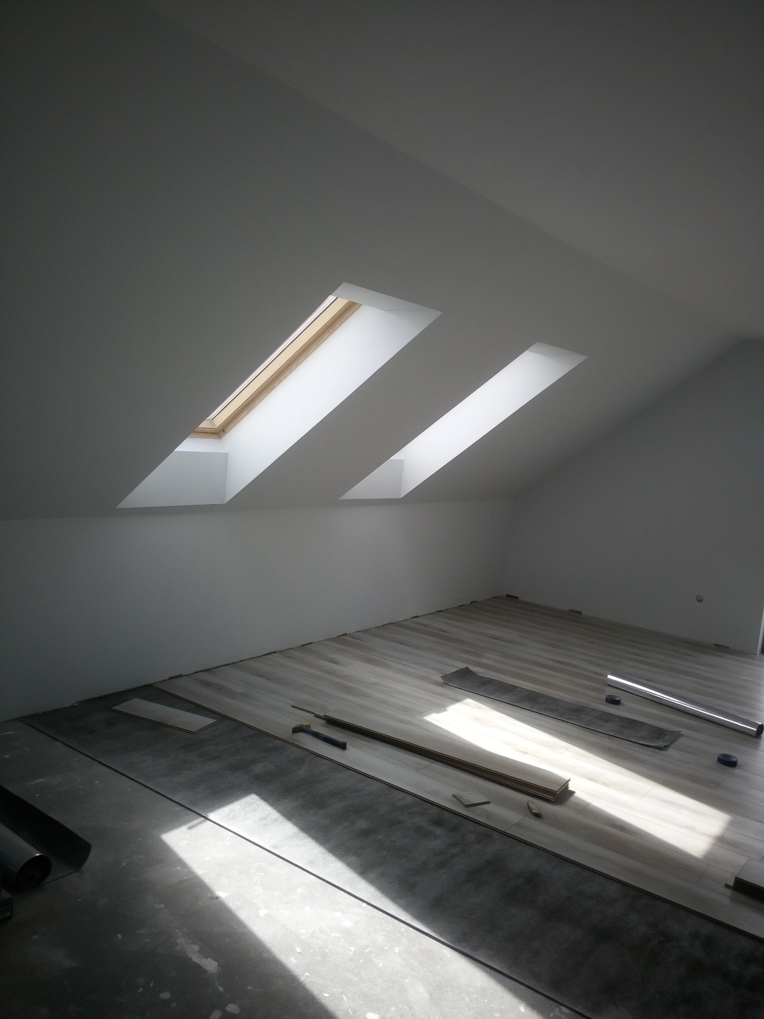 Montaż paneli podłogowych w jasnym pokoju na poddaszu z dwoma oknami dachowymi, widoczne narzędzia i materiały budowlane.