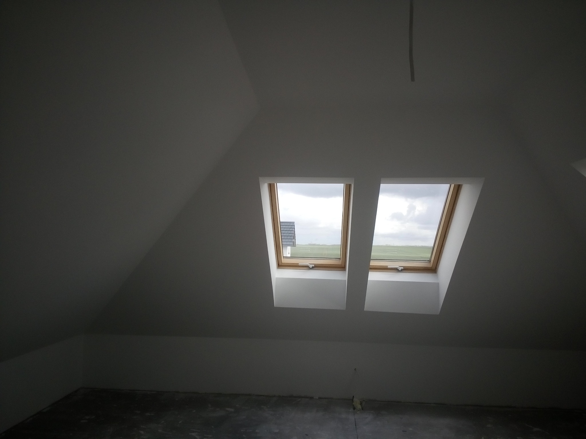Poddasze po wykonaniu gładzi, widoczne dwa okna dachowe z drewnianymi ramami, szare ściany i podłoga w stanie surowym.